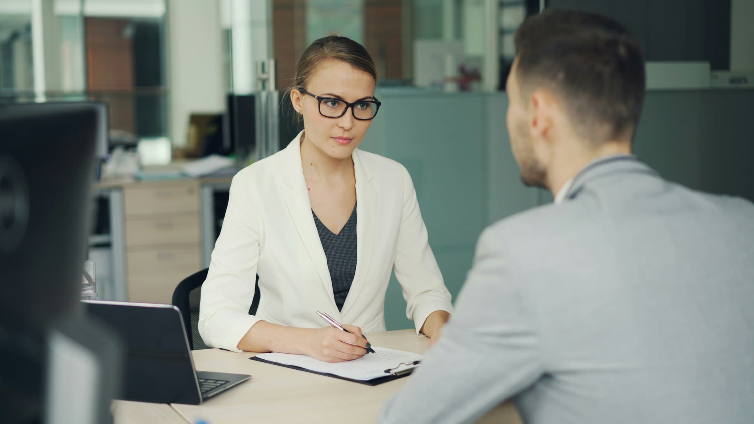 A woman in glasses takes notes while questioning a man across an office desk.
