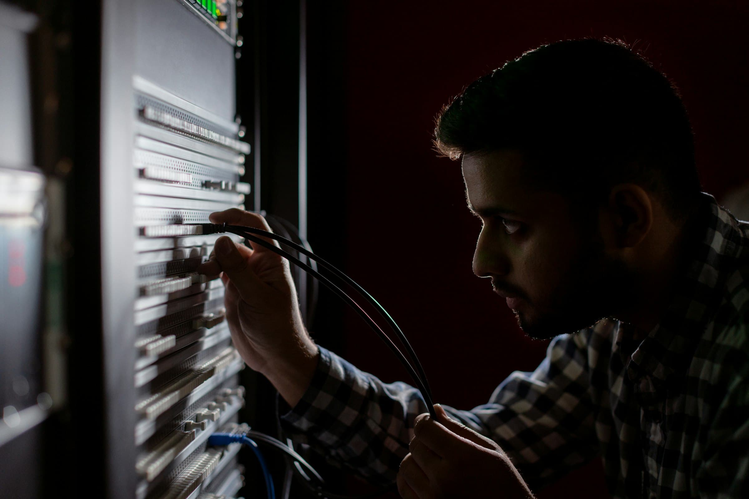 A man in a dark room carefully plugs a cable into a server rack.